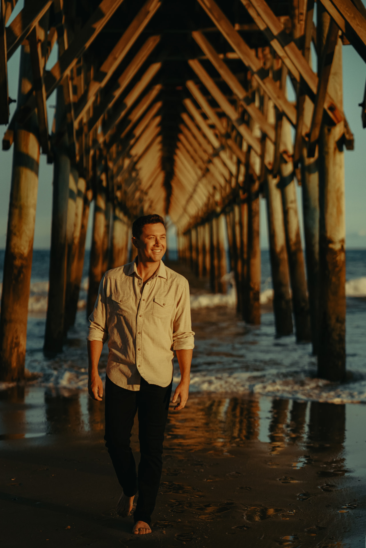 Scotty McCreery walking barefoot on the beach beneath a pier.